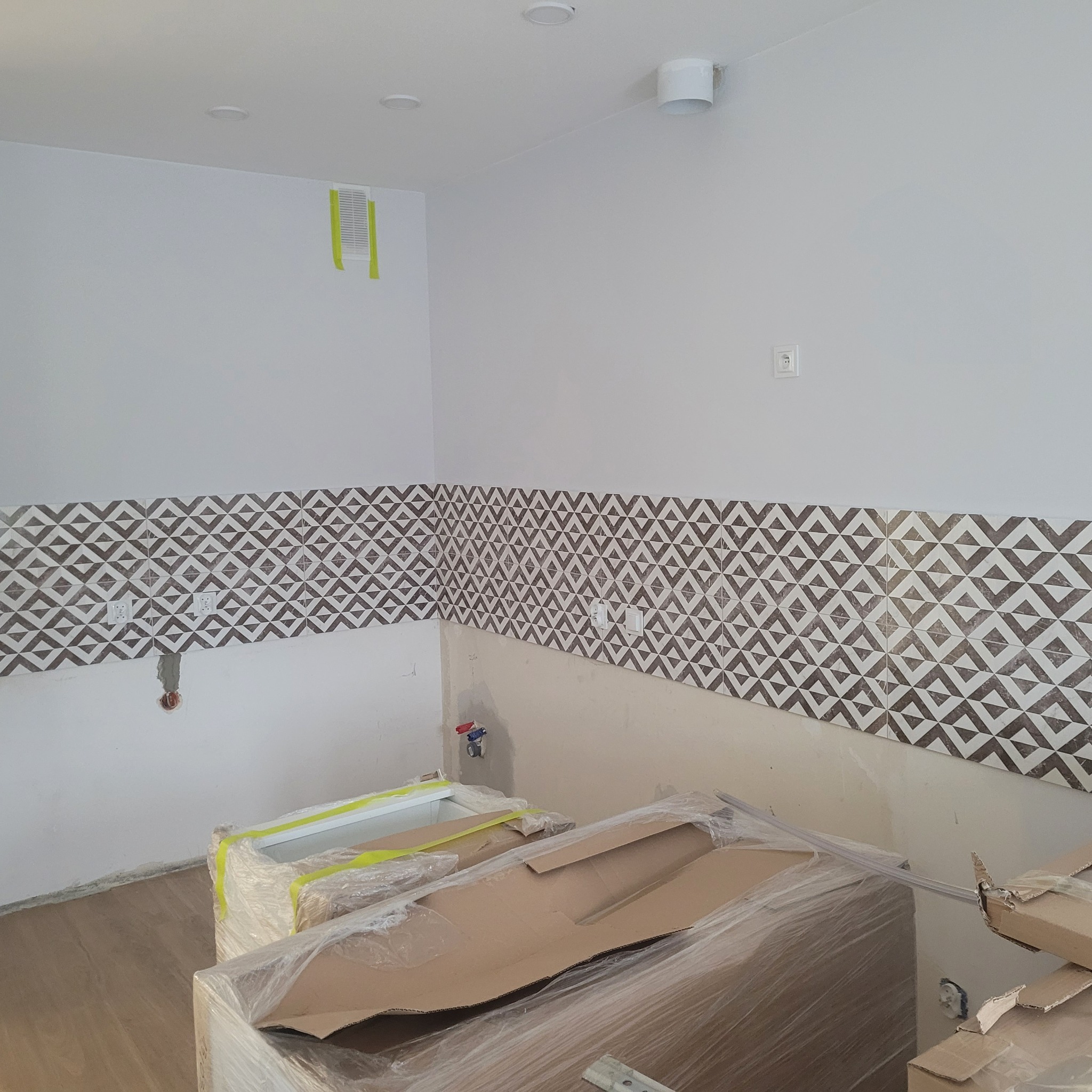 Fragment kuchni w trakcie prac wykończeniowych: częściowo położone płytki dekoracyjne z geometrycznym wzorem, zabezpieczone folią meble kuchenne, widoczne punkty instalacji elektrycznej i wodnej.