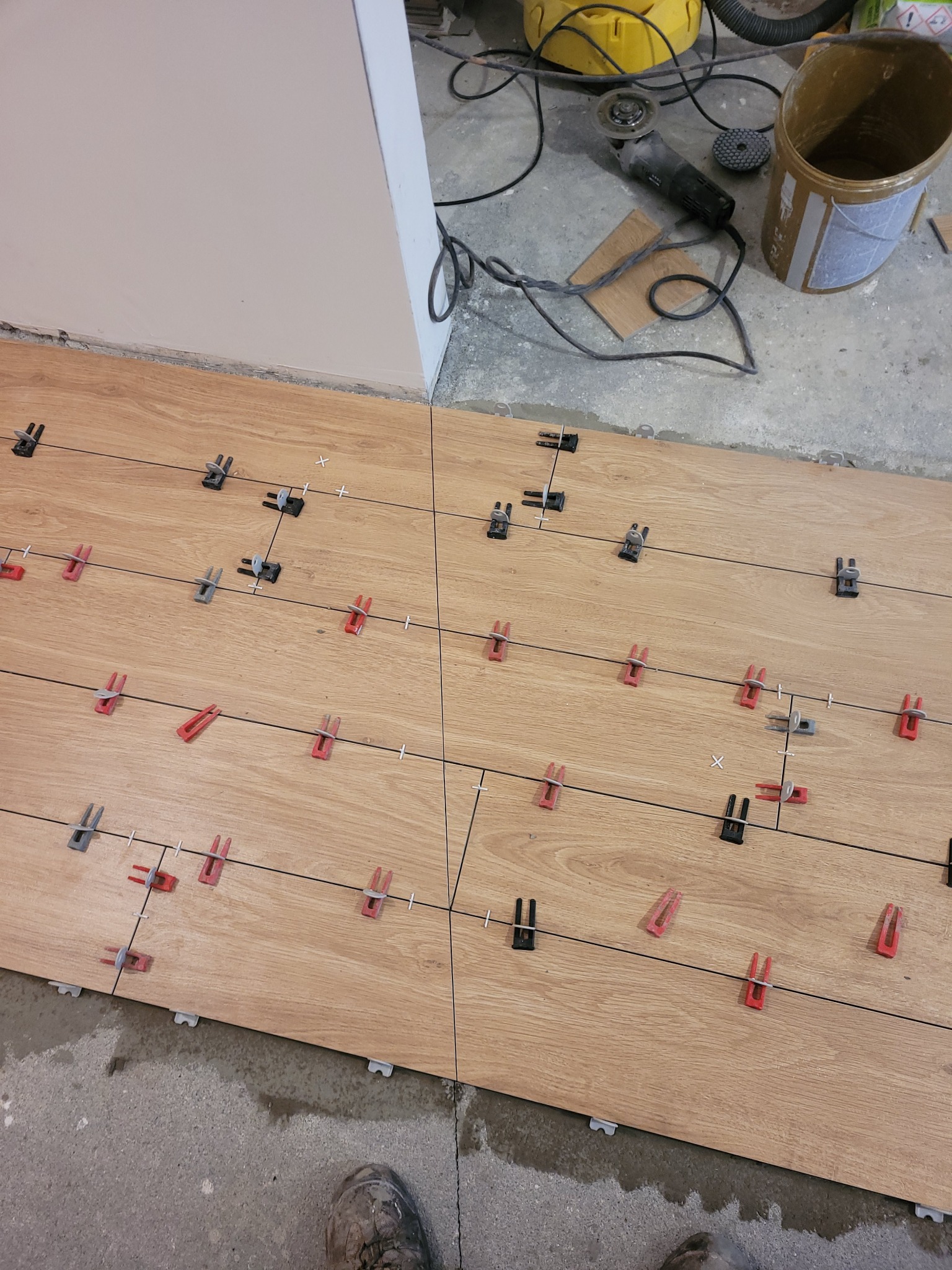 Układanie płytek podłogowych imitujących drewno, z użyciem klipsów poziomujących, widoczny fragment buta roboczego i narzędzia w tle.