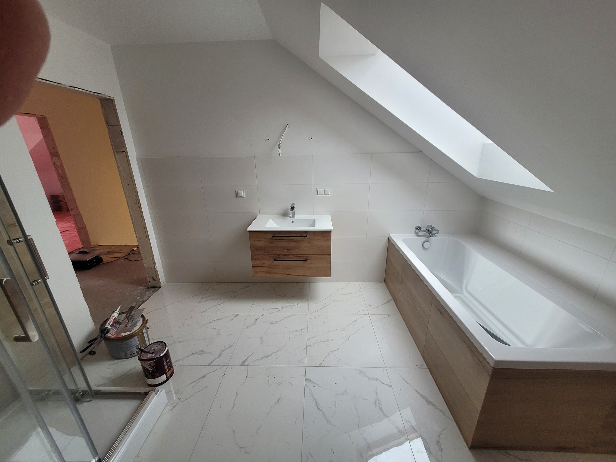 Ukończona łazienka na poddaszu ze skośnym oknem dachowym, białe płytki na ścianach i podłodze z marmurowym wzorem, umywalka z szafką podwieszaną w kolorze drewna, wanna obudowana drewnopodobnymi...