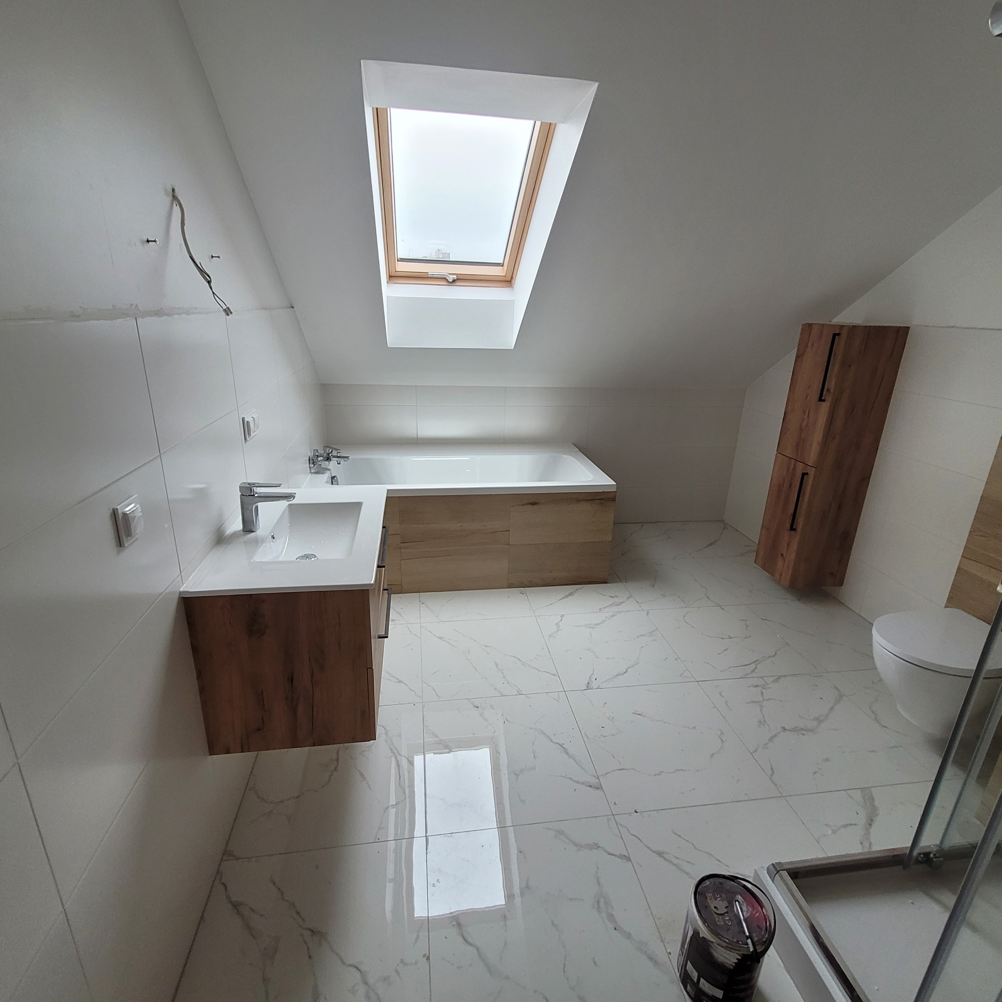 Wykończona łazienka na poddaszu z oknem dachowym: białe płytki marmurowe, drewniana szafka pod umywalką z białym blatem, zabudowana wanna, drewniana szafka wisząca. Na podłodze wiadro z farbą.