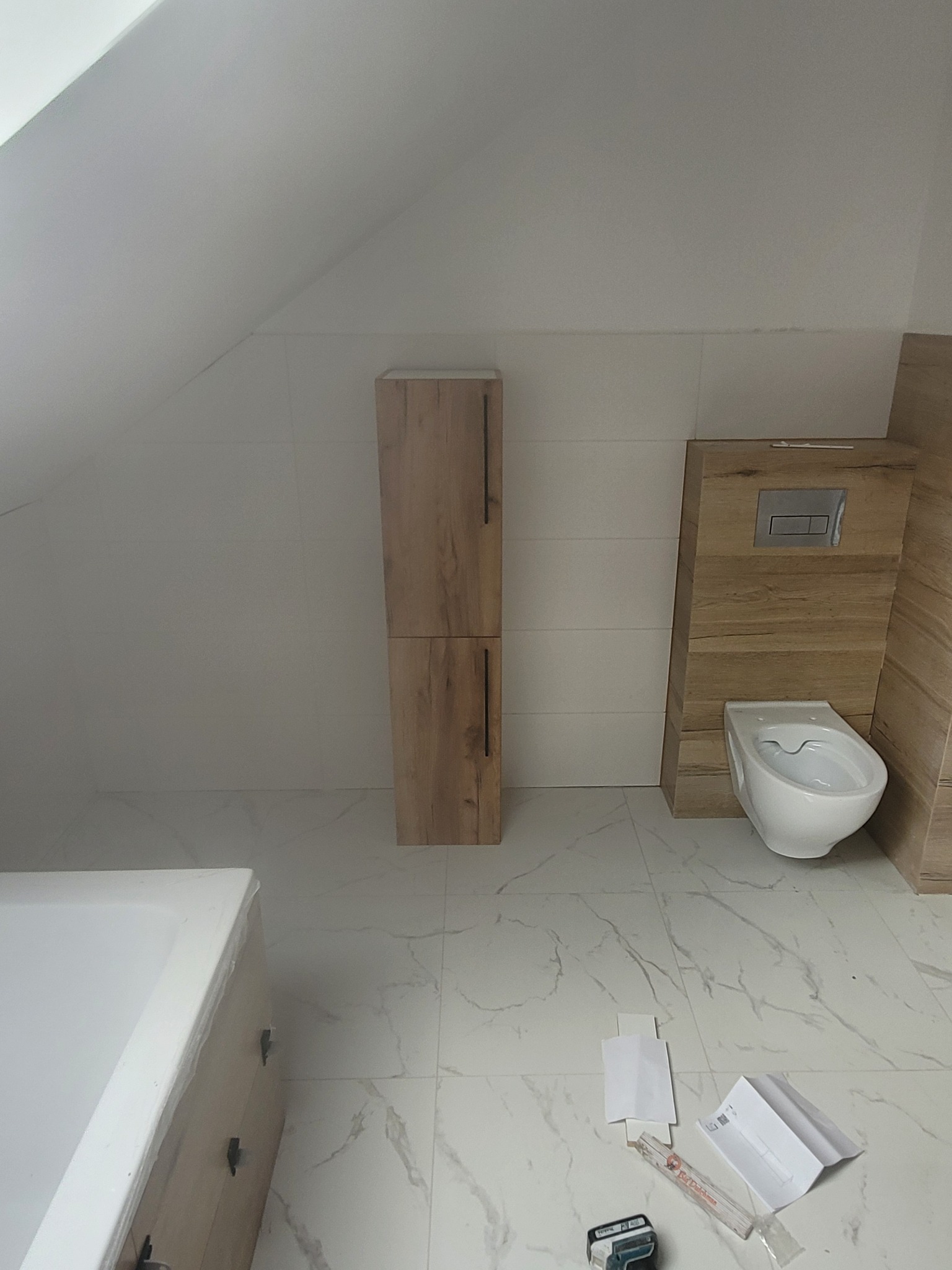 Ukośna łazienka z białą armaturą i drewnopodobnymi szafkami; widoczne narzędzia i resztki po montażu na marmurowej podłodze.