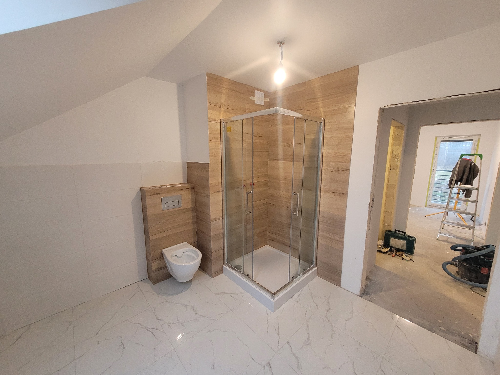 Nowoczesna łazienka w trakcie wykańczania: kabina prysznicowa z przezroczystego szkła, podwieszana toaleta z drewnopodobną obudową, białe płytki marmurowe na podłodze i ścianach, widoczne narzędzia...