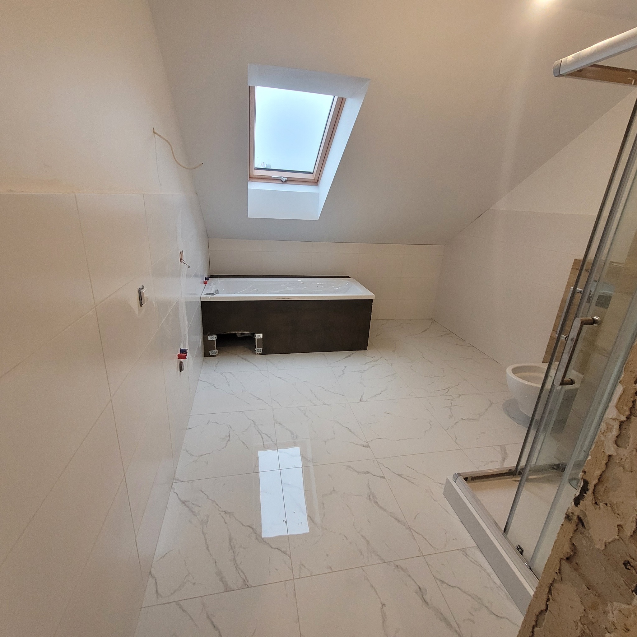 Łazienka na poddaszu w trakcie prac wykończeniowych: białe płytki z marmurowym wzorem na podłodze, wanna z obudową w kolorze ciemnego brązu, częściowo zamontowana kabina prysznicowa, widoczne kable...