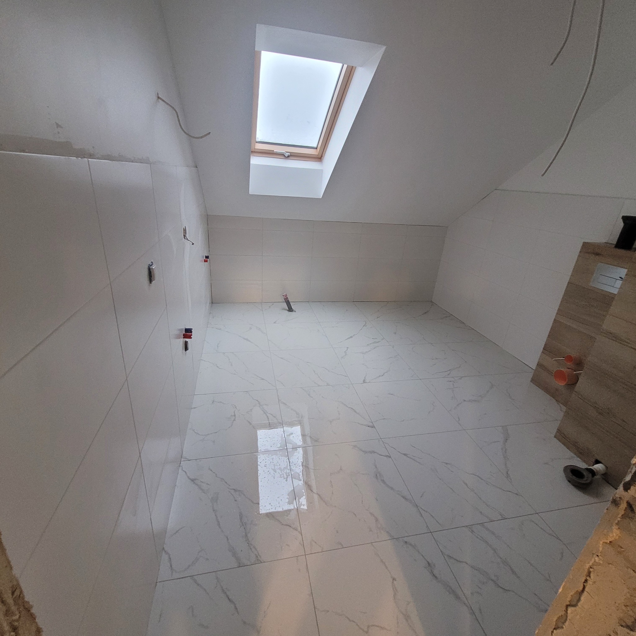 Łazienka na poddaszu w trakcie remontu: białe płytki imitujące marmur na podłodze i ścianach, okno dachowe, wystające kable elektryczne, widoczne mocowania do armatury.