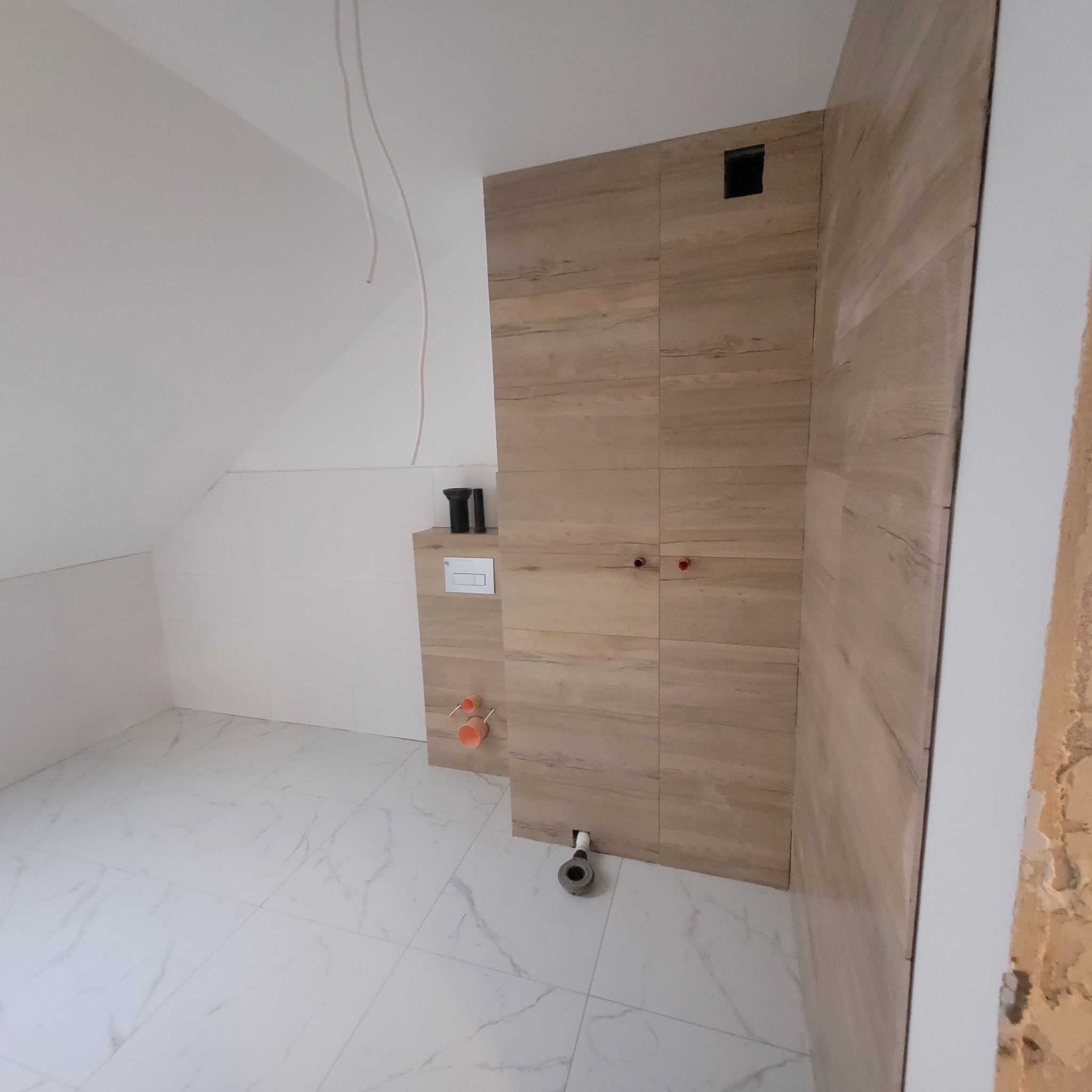 Wnętrze łazienki w trakcie prac wykończeniowych; widoczne płytki imitujące drewno na ścianie, białe płytki podłogowe z marmurkowym wzorem, wystające rury instalacyjne i przewody elektryczne.