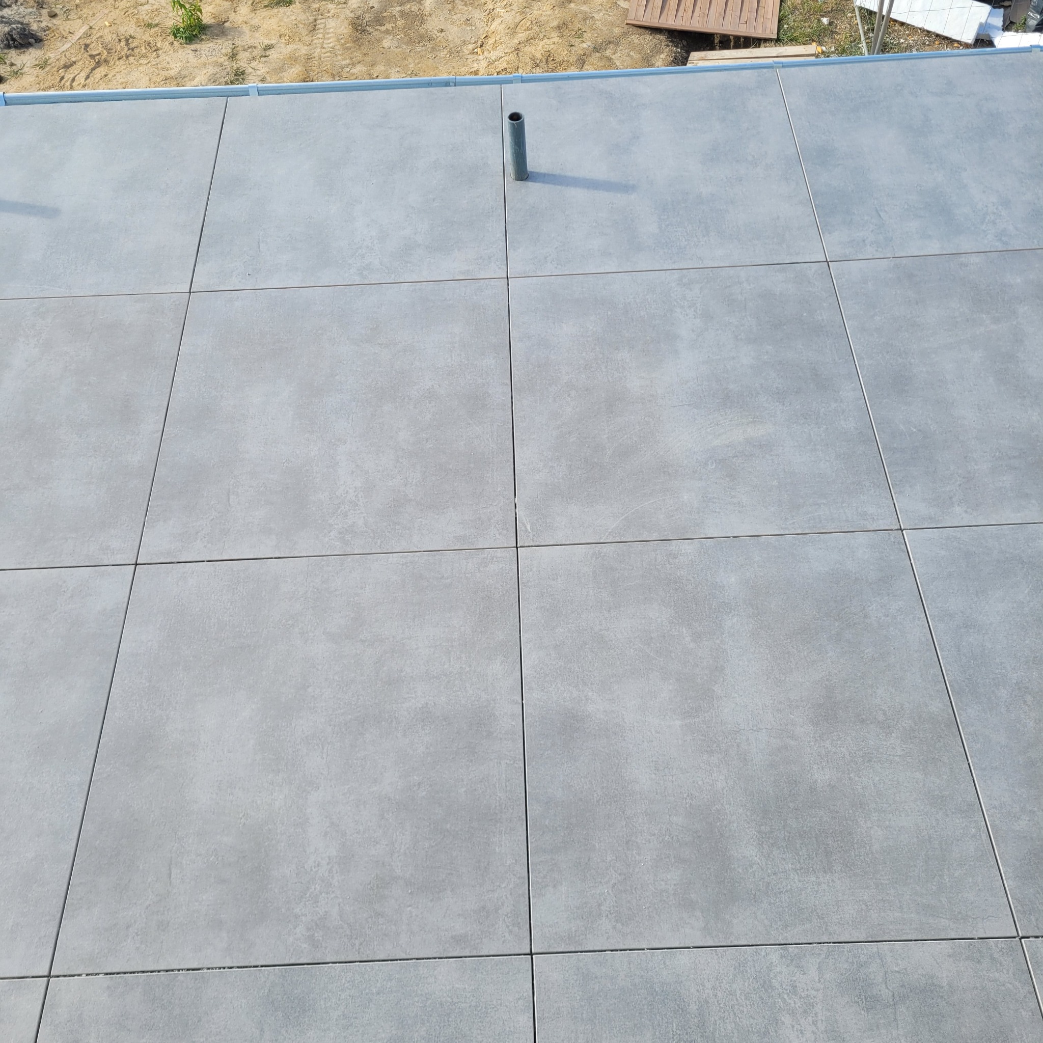 Szare, kwadratowe płytki ułożone na podłodze lub tarasie, widoczna rura wystająca z powierzchni i fragment elewacji.