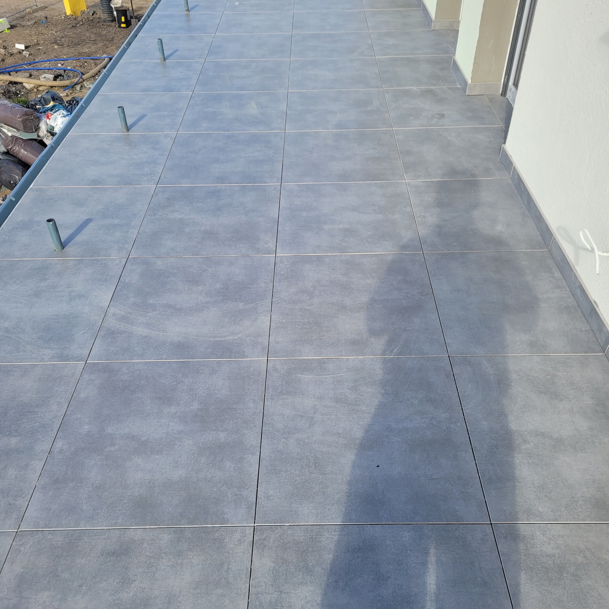 Ułożona szara terakota na tarasie, widoczne rury wystające z podłoża przygotowane pod montaż balustrady, w tle fragment ściany budynku.