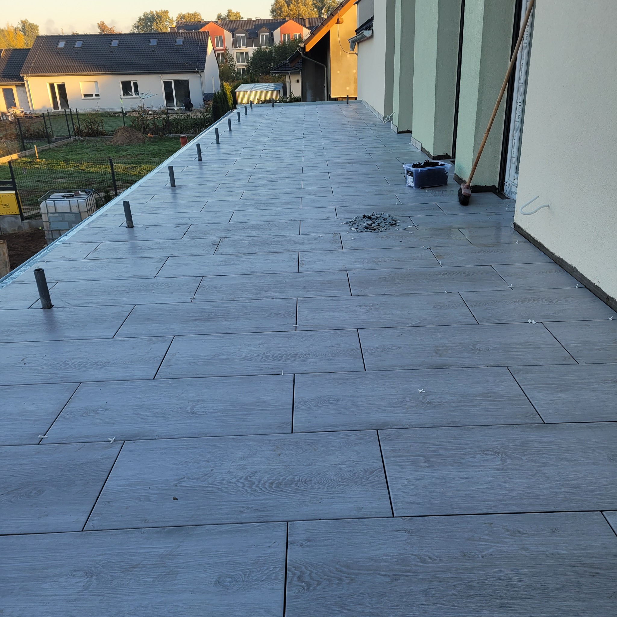 Układanie szarych płytek imitujących drewno na balkonie, widoczne krzyżyki dystansowe, rury do balustrady i narzędzia.