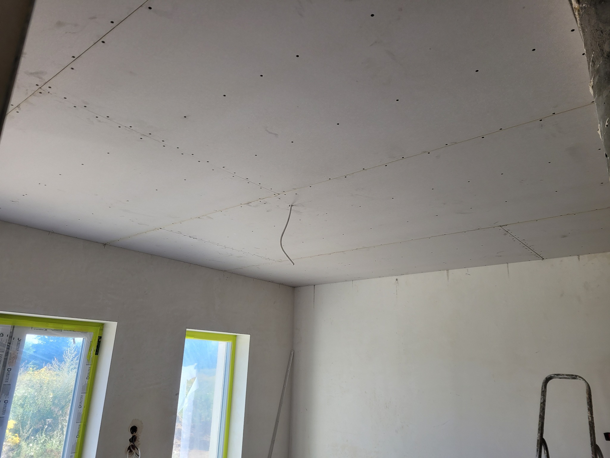 Montaż płyt gipsowo-kartonowych na suficie w trakcie remontu, widoczne spoiny i wkręty, dwa okna z zielonymi ramami w tle, drabina oparta o ścianę.