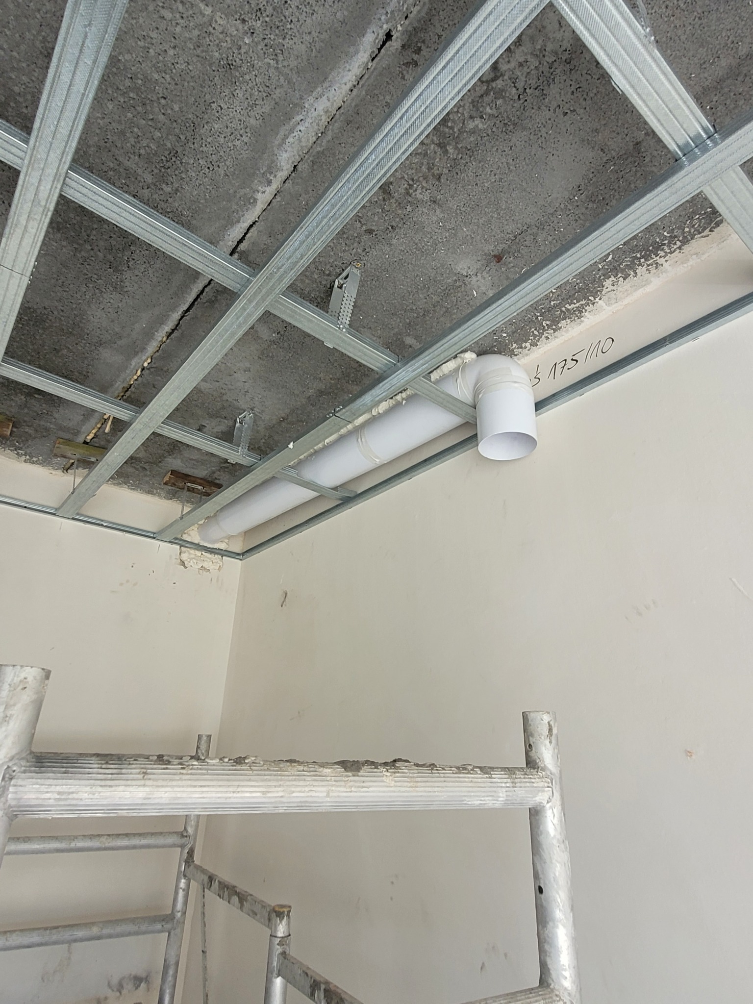 Montaż instalacji wentylacyjnej z rur PCV wewnątrz metalowej konstrukcji sufitu podwieszanego, widoczne rusztowanie malarskie.