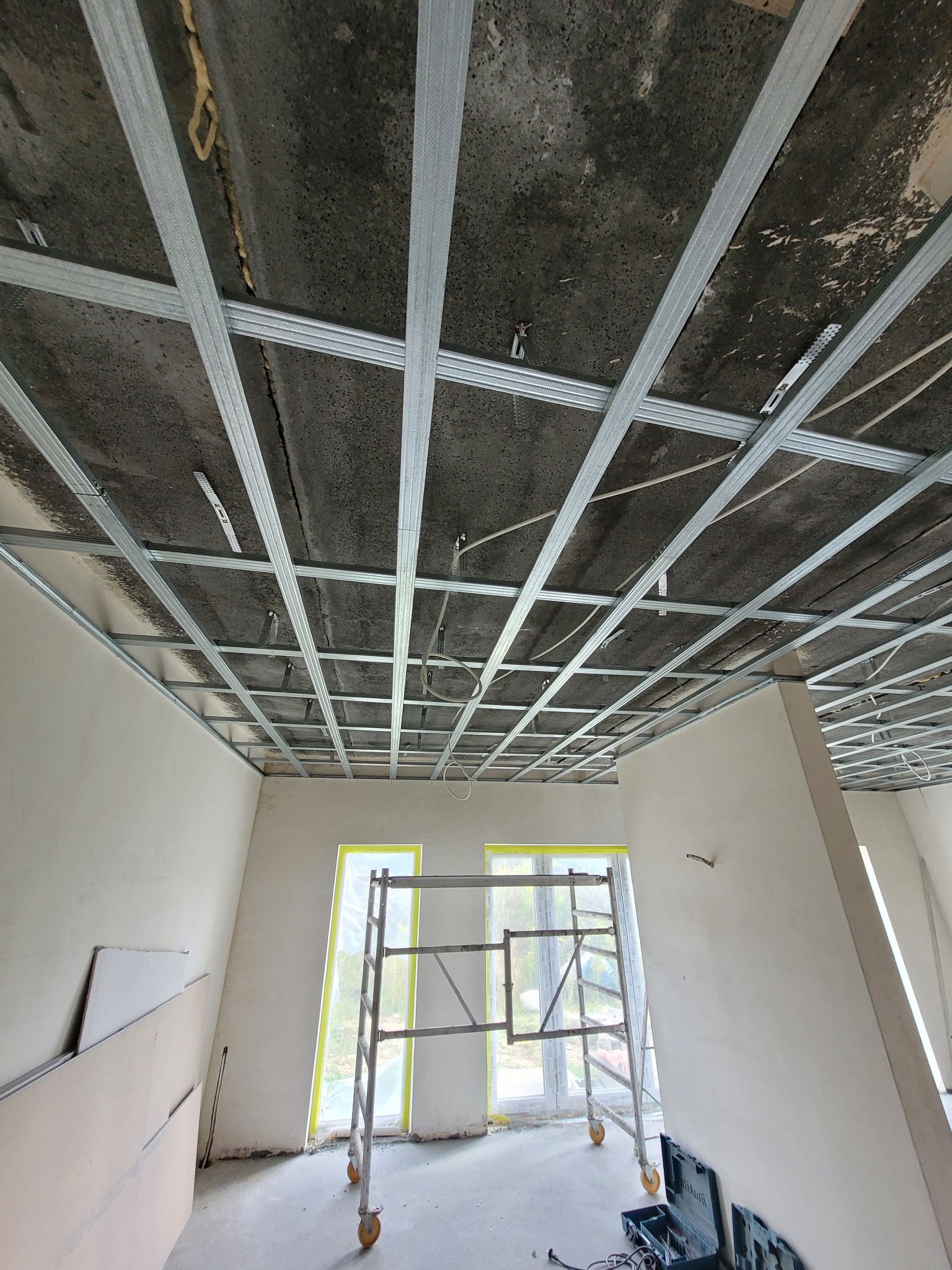 Montaż metalowej konstrukcji sufitu podwieszanego w pomieszczeniu z surowym betonowym stropem i zabezpieczonymi folią oknami, widoczny także rusztowanie i płyty gipsowo-kartonowe.