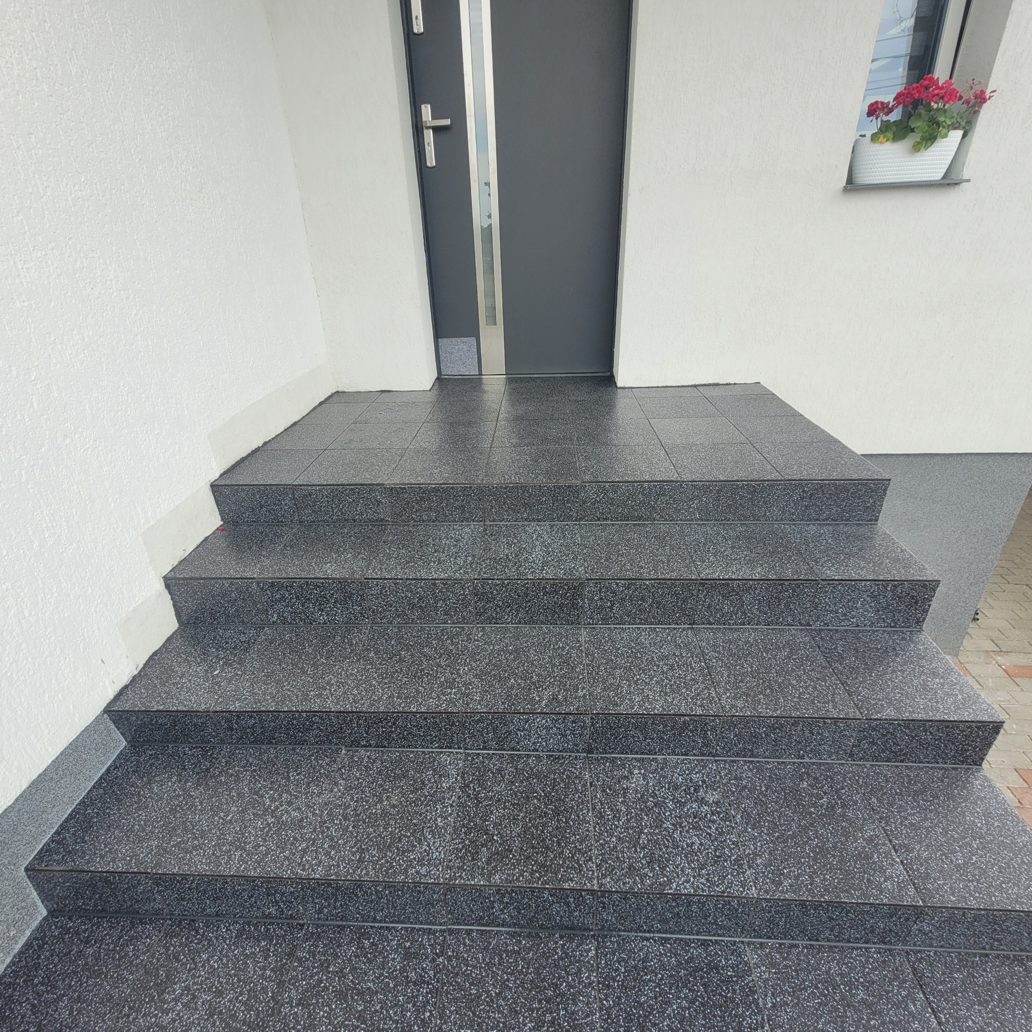 Zewnętrzne schody wejściowe wyłożone ciemnymi, błyszczącymi płytkami z delikatnym wzorem, prowadzące do szarych drzwi wejściowych z szybą i kwiatów w doniczce na parapecie okna.
