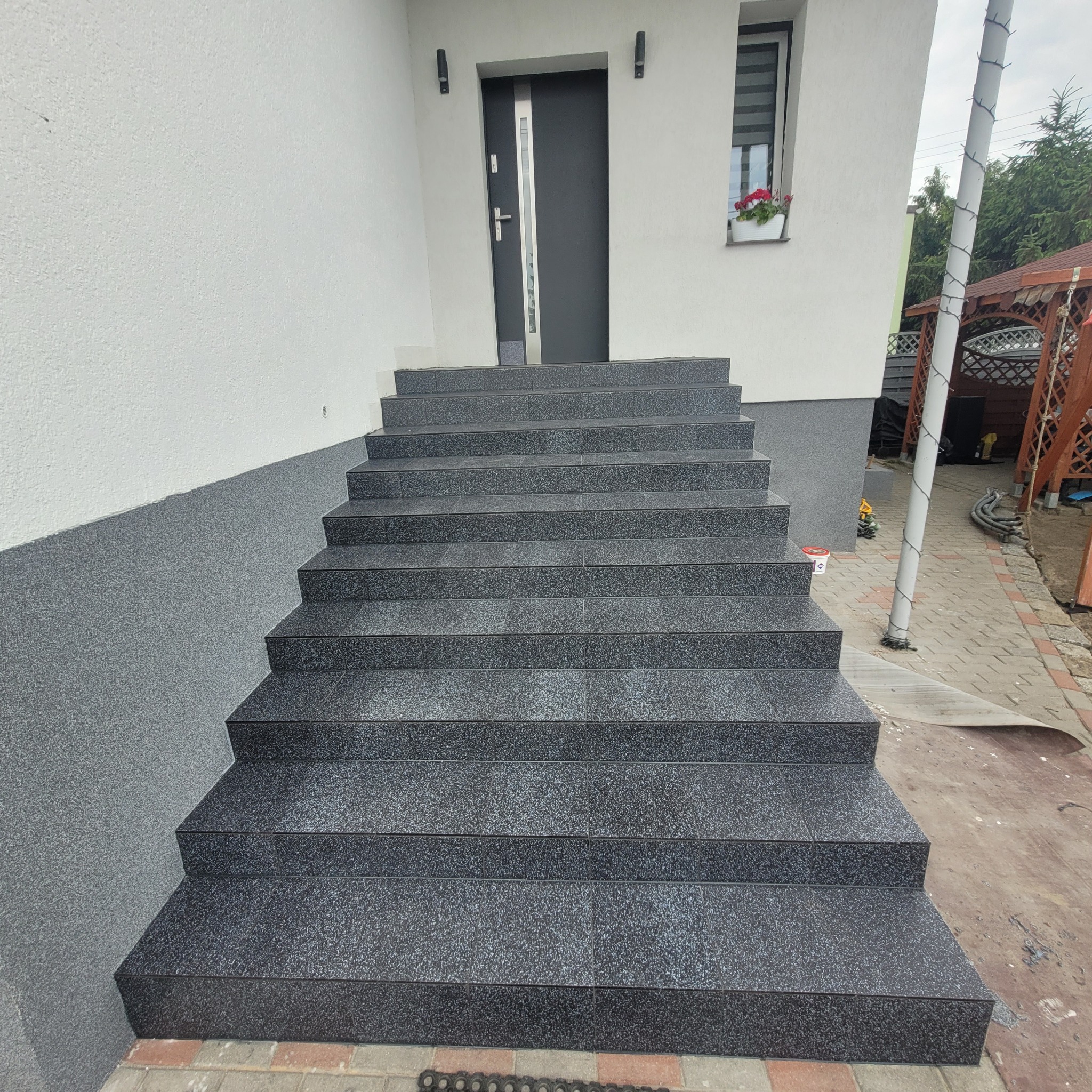 Zewnętrzne schody wejściowe wyłożone ciemnoszarymi płytkami z drobnym wzorem, prowadzące do nowoczesnych drzwi wejściowych z pionowym przeszkleniem, ściana budynku z jasnym tynkiem strukturalnym...