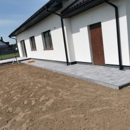 Firma Ogólnobudowlana DAM Damian Nowosad - Fragment nowo wyłożonej, szarej kostki brukowej przy białym budynku jednorodzinnym, widoczny laser niwelacyjny i przygotowany teren pod dalsze prace.