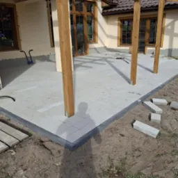 Szara nawierzchnia z prostokątnych płyt ułożona wokół drewnianych słupów z widocznymi niedociągnięciami na krawędziach i stertą kostek obok, na tle domu z brązowymi oknami.