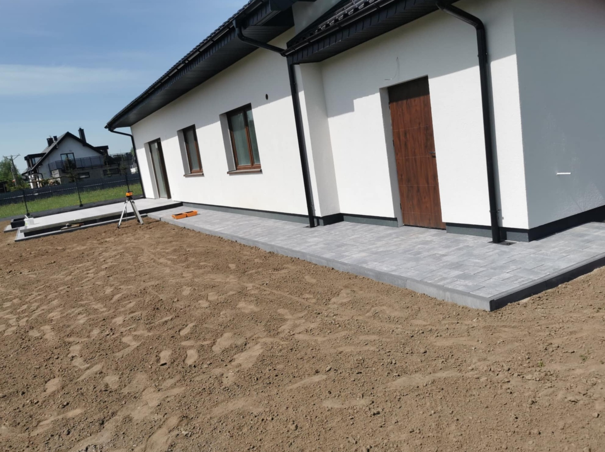 Fragment nowo wyłożonej, szarej kostki brukowej przy białym budynku jednorodzinnym, widoczny laser niwelacyjny i przygotowany teren pod dalsze prace.