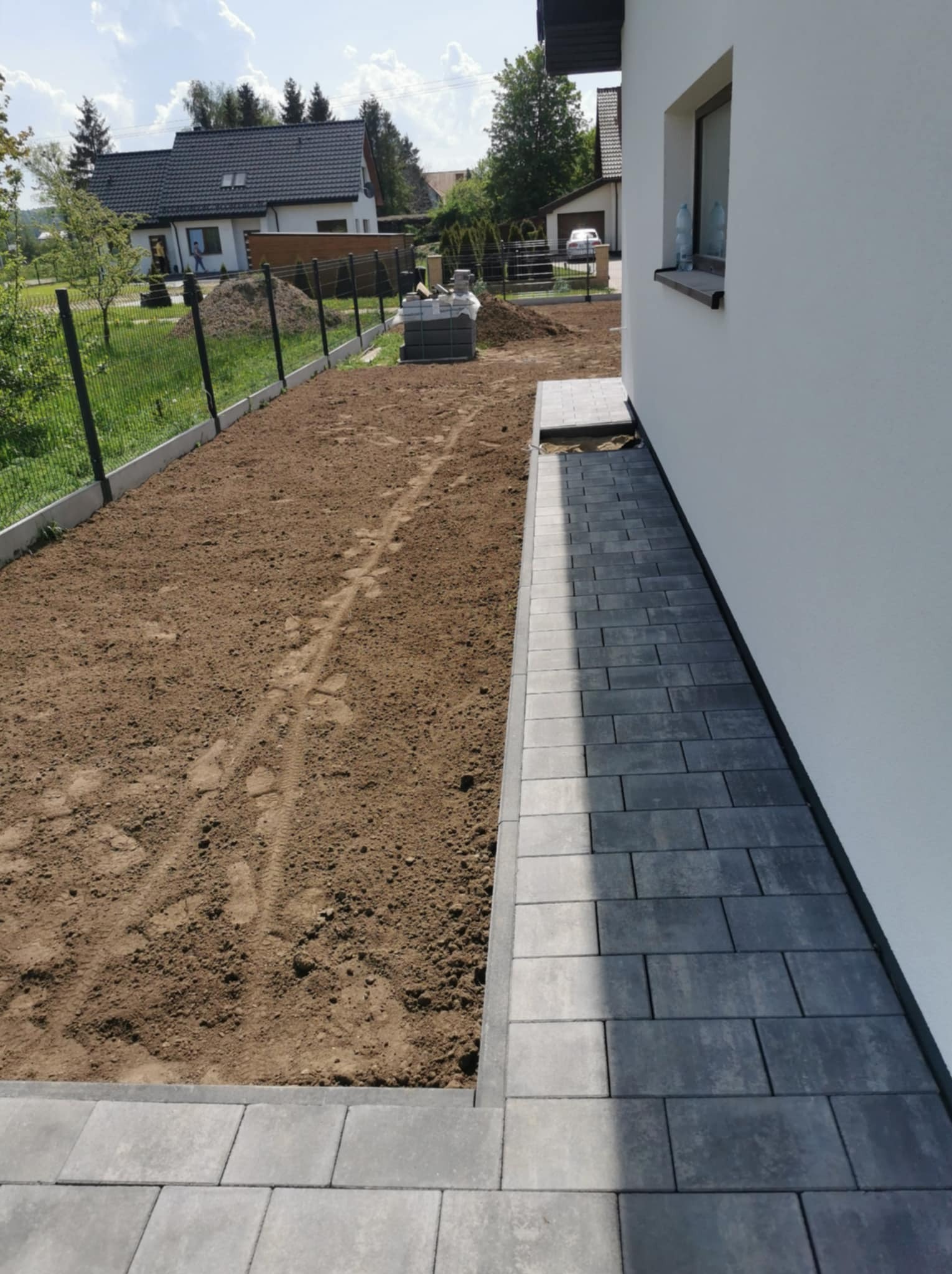 Częściowo ułożona, ciemnoszara kostka brukowa przy białej ścianie domu w Łęczycach, widoczne przygotowane podłoże i paleta z kostką w tle.