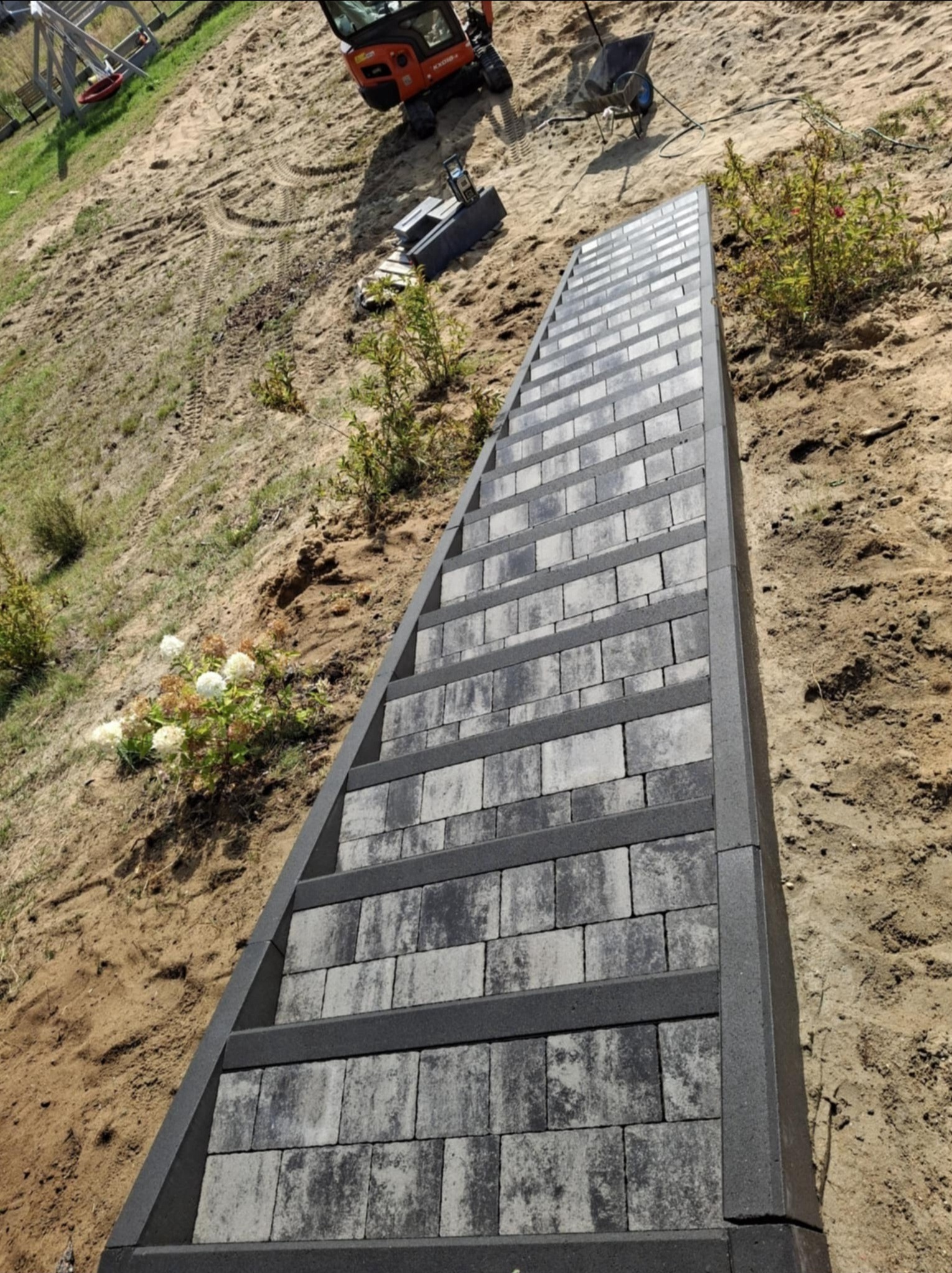 Schody zewnętrzne z kostki brukowej w odcieniach szarości, prowadzące w górę po piaszczystym zboczu, z widocznym minikoparką i taczką w tle.