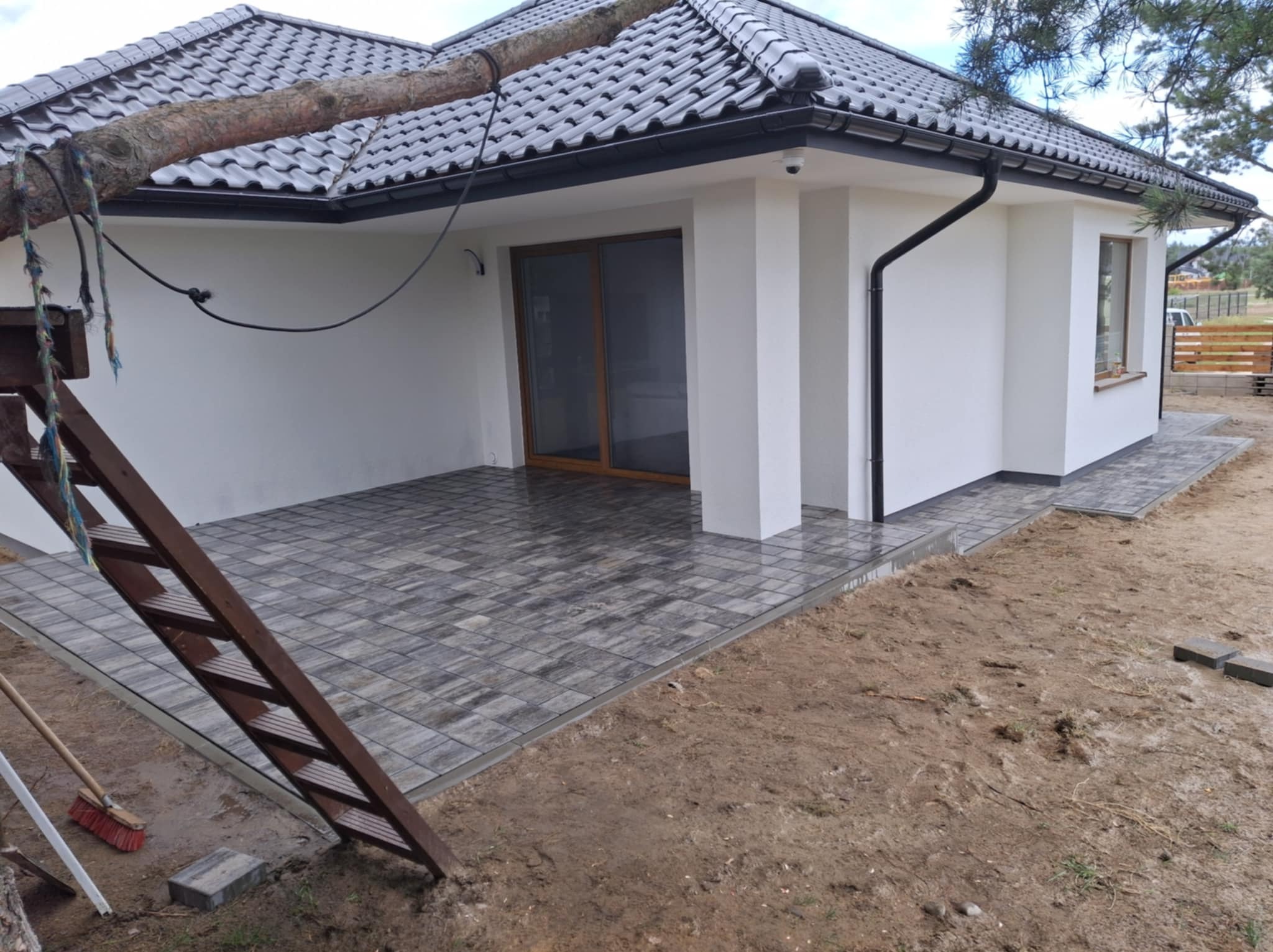 Nowoczesny taras z szarej kostki brukowej ułożonej przy białym domu z ciemną dachówką, widok na schody i przygotowany teren.