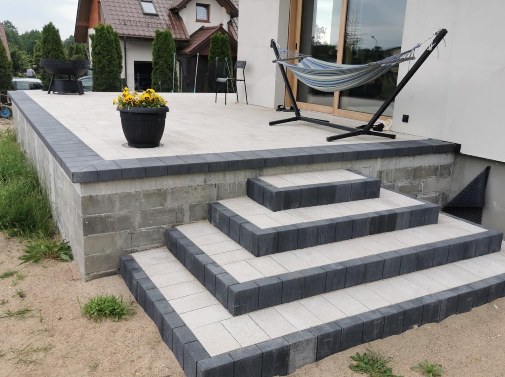 Taras z kostki brukowej w odcieniach szarości i beżu z czterostopniowymi schodami, donicą z żółtymi kwiatami i hamakiem w tle.