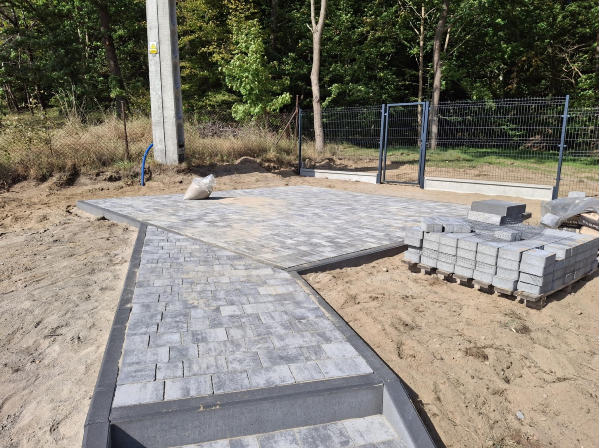 Wykonana z szarej kostki brukowej ścieżka i plac przed domem, z widocznymi paletami kostki brukowej i workiem piasku, w tle ogrodzenie z siatki.