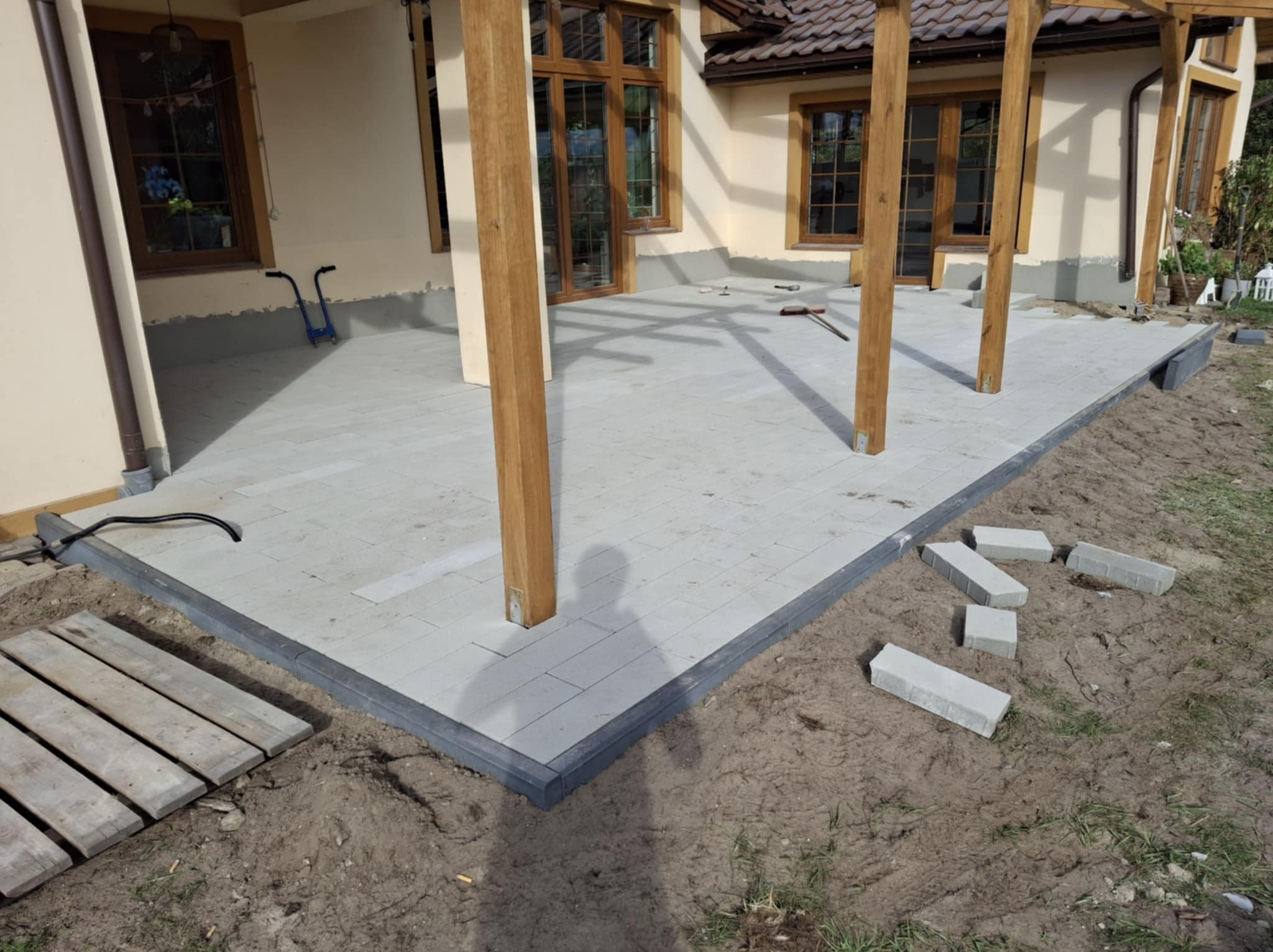 Szara nawierzchnia z prostokątnych płyt ułożona wokół drewnianych słupów z widocznymi niedociągnięciami na krawędziach i stertą kostek obok, na tle domu z brązowymi oknami.