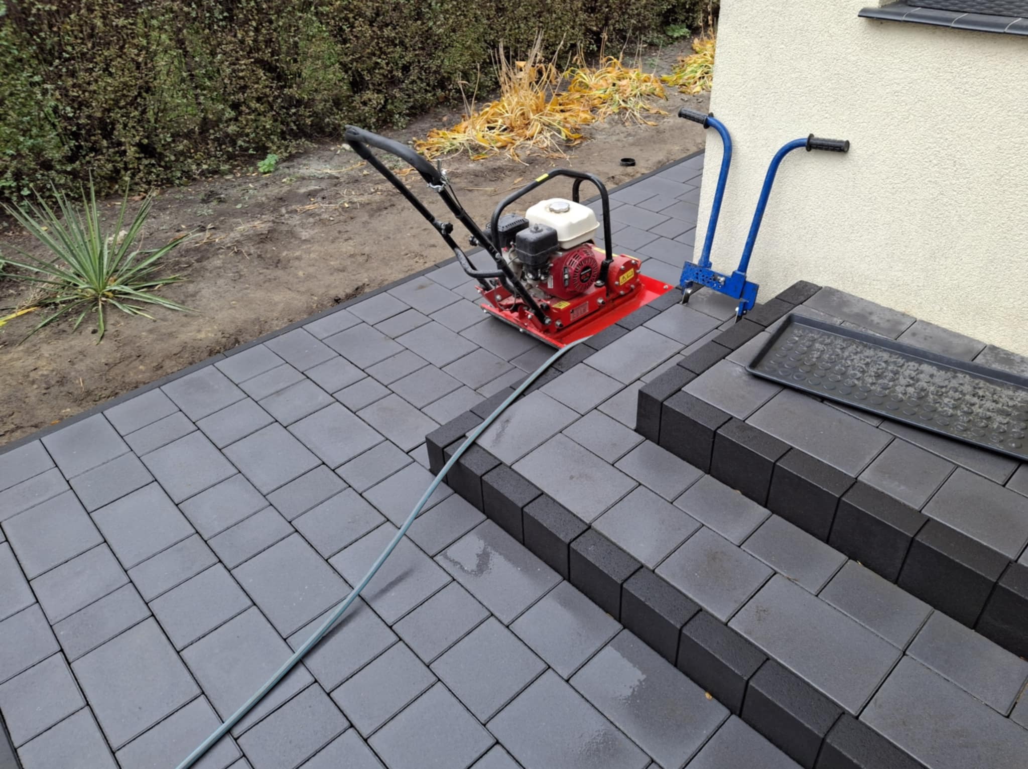 Częściowo ułożona szara kostka brukowa z czerwoną zagęszczarką i niebieskimi narzędziami do układania kostki, widoczne schody z kostki i fragment trawnika z roślinnością.
