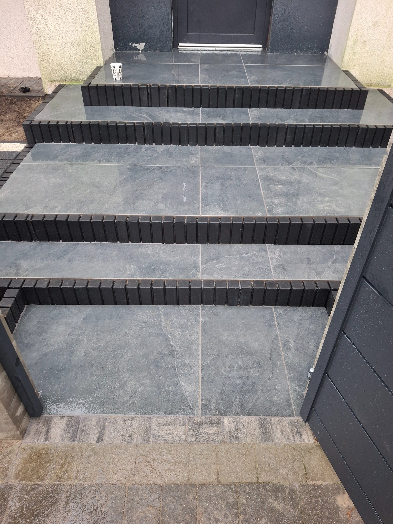 Schody wejściowe wyłożone szarą kostką z czarnymi obrzeżami, widok z góry, częściowo widoczne drzwi wejściowe i fragment ogrodzenia. Na schodach stoi kubek.