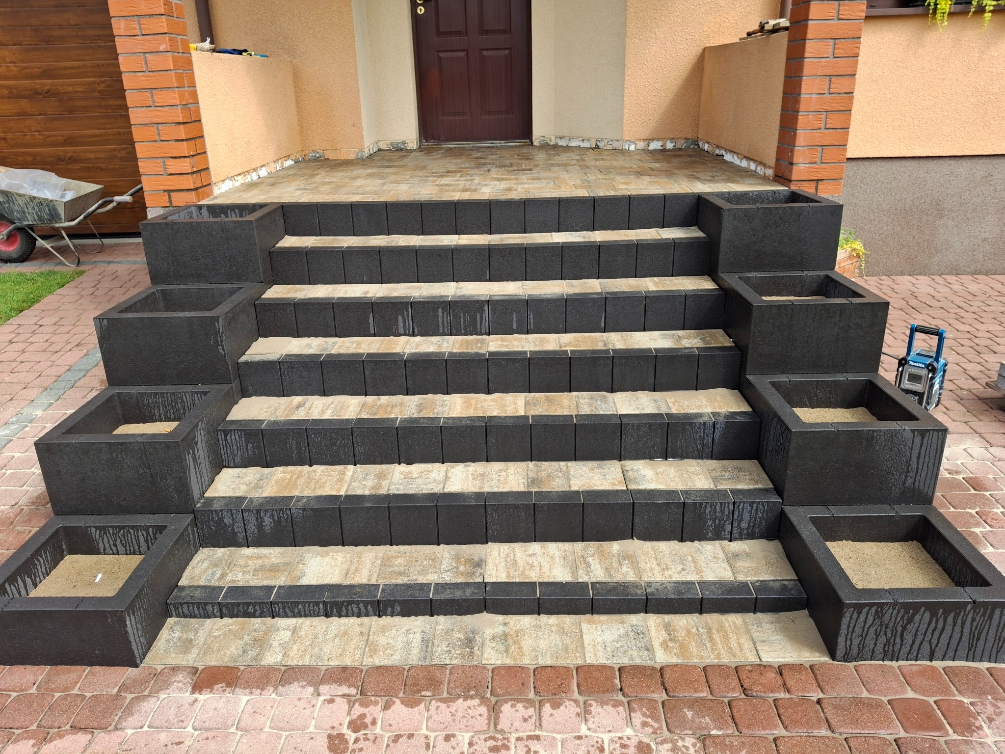 Schody wejściowe z kostki brukowej w dwóch odcieniach, z donicami wypełnionymi piaskiem. Czerwona kostka na obrzeżach. Widoczny wózek i narzędzia.