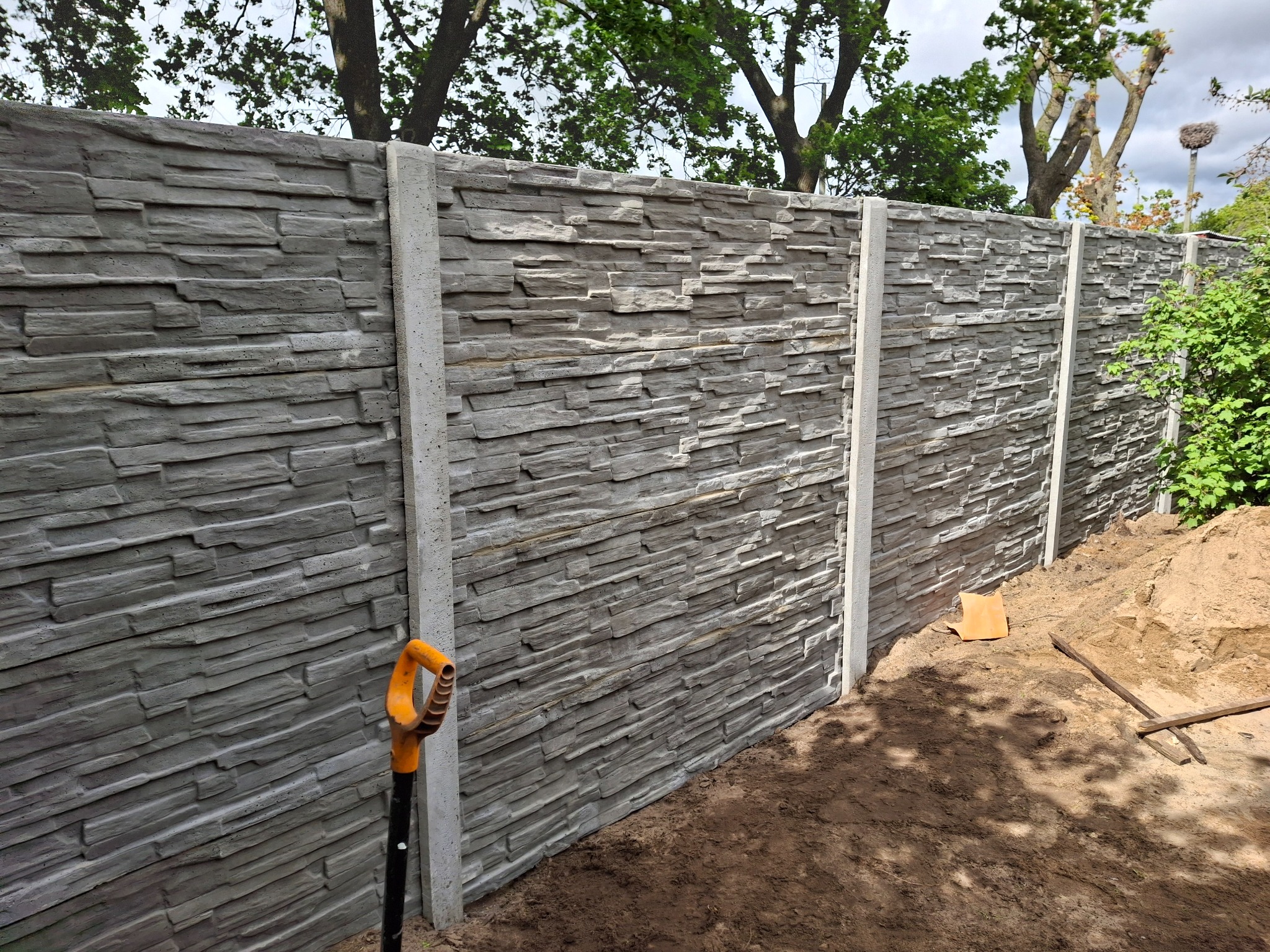 Szary, panelowy płot z imitacją kamienia, wsparty na betonowych słupkach. W tle drzewa i niebo. Przy płocie stoi szpadel z pomarańczową rączką i leży ziemia.