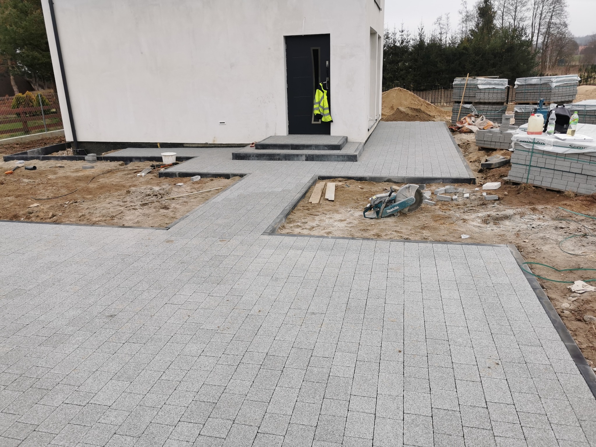 Jasnoszara kostka brukowa ułożona na podjeździe i ścieżce prowadzącej do domu. Widoczny fragment budynku z czarnymi drzwiami i sterta kostki brukowej w tle.