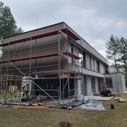 SzwagrBud - Nowoczesny dom w trakcie wykończenia elewacji, z widocznym rusztowaniem, elementami ocieplenia i pracownikiem na dachu, otoczony drzewami i trawnikiem.