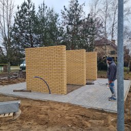 SzwagrBud - Wznoszenie ścianek działowych z żółtej cegły klinkierowej na zewnątrz budynku, widoczni dwaj pracownicy w trakcie prac wykończeniowych, teren przygotowany pod dalsze prace.