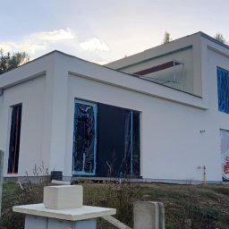 SzwagrBud - Nowoczesny, biały dom w Białymstoku w trakcie prac elewacyjnych, okna zabezpieczone folią, widoczne rusztowania i materiały budowlane na posesji.