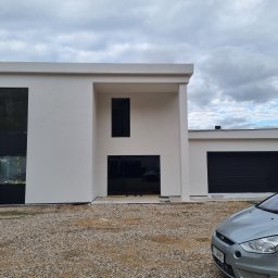 SzwagrBud - Nowoczesny dom z białymi ścianami i czarnymi oknami, pracownik na rusztowaniu maluje elewację w pobliżu okna, widoczny samochód na żwirowym podłożu.