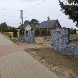 SzwagrBud - Wznoszenie ogrodzenia z szarej, betonowej kostki modułowej w sąsiedztwie chodnika z kostki brukowej i drogi, widoczne domy w tle.