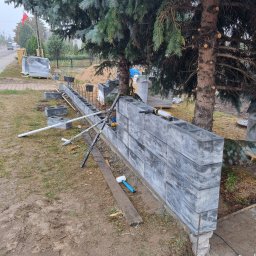 SzwagrBud - Wznoszenie muru z szarych betonowych bloczków w ogrodzie, widoczne narzędzia: poziomica, młotek gumowy, pistolet do kleju, w tle drzewa iglaste i palety z bloczkami.