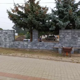 Budowa ogrodzenia z szarych, betonowych bloczków; widoczny murarz przy pracy, taczka i stosy materiałów budowlanych w tle.