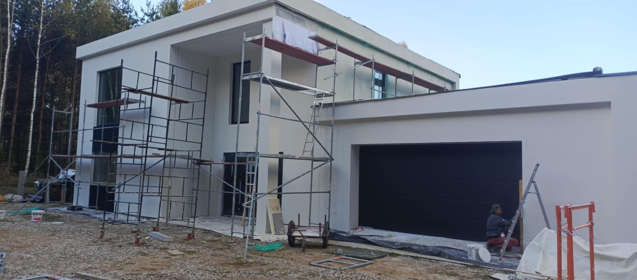 Nowoczesny dom jednorodzinny w trakcie prac elewacyjnych, widoczne rusztowania i pracownik malujący fragment ściany przy bramie garażowej.