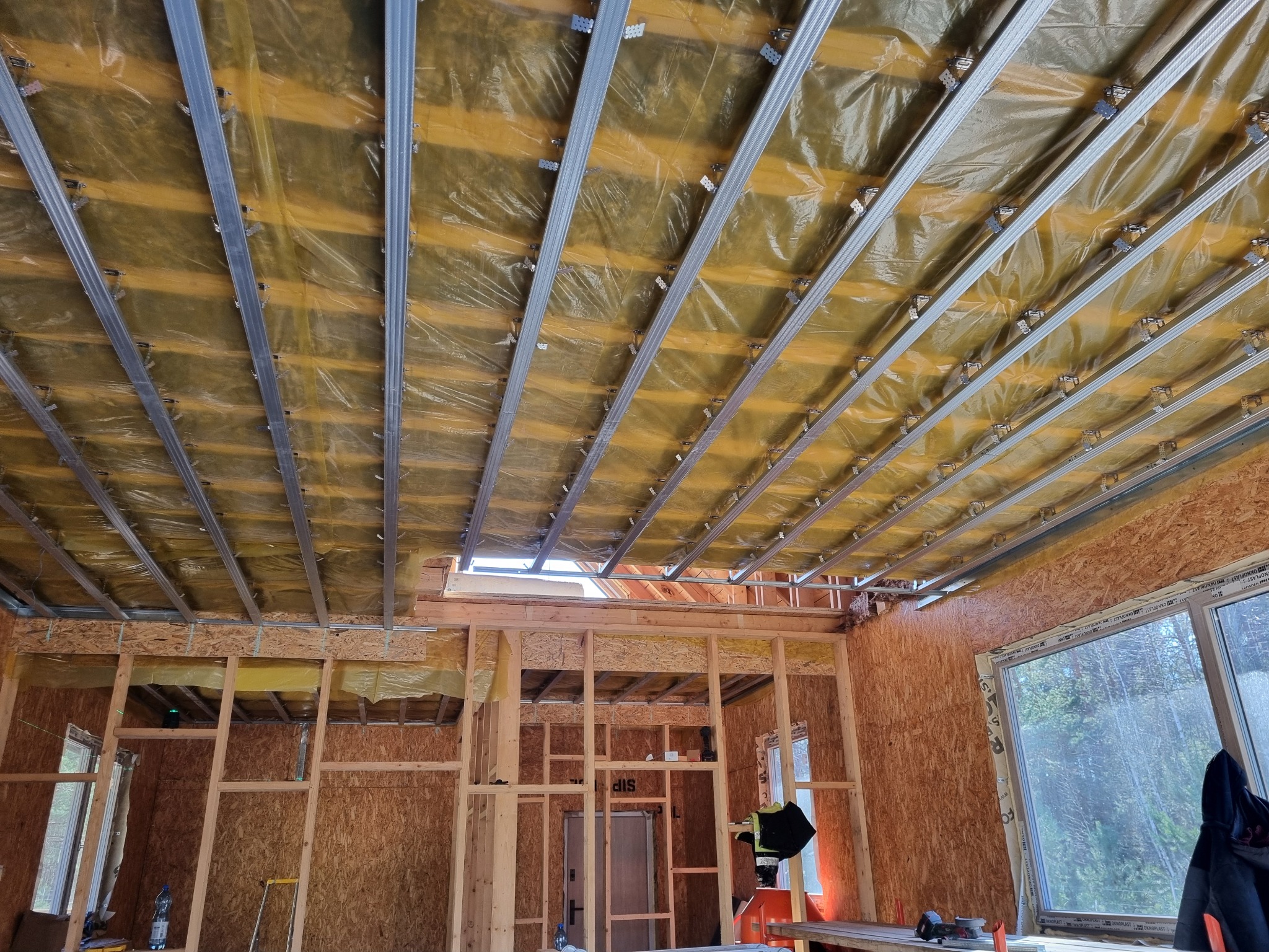 Wnętrze domu w trakcie budowy, widoczne drewniane szkielety ścian obłożone płytami OSB, folia paroizolacyjna na suficie i metalowe profile sufitowe