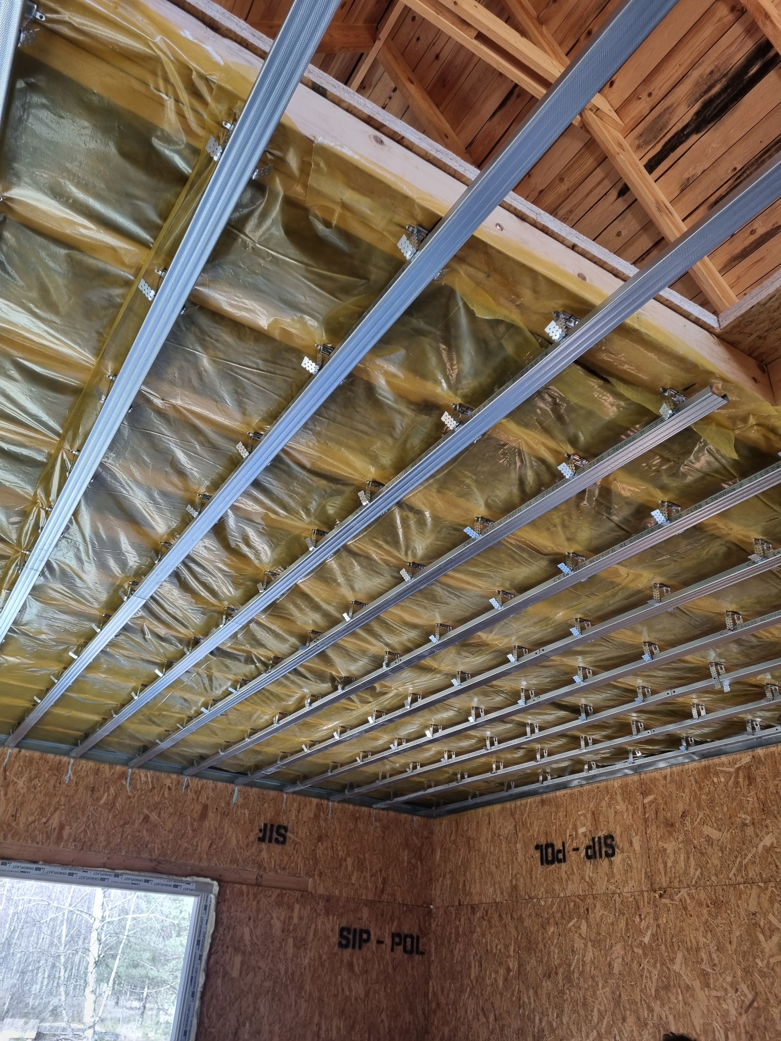 Wnętrze budynku w trakcie prac wykończeniowych: widoczna izolacja folią paroizolacyjną pod drewnianym stropem i metalowy stelaż do montażu sufitu podwieszanego; ściany obłożone płytami OSB...