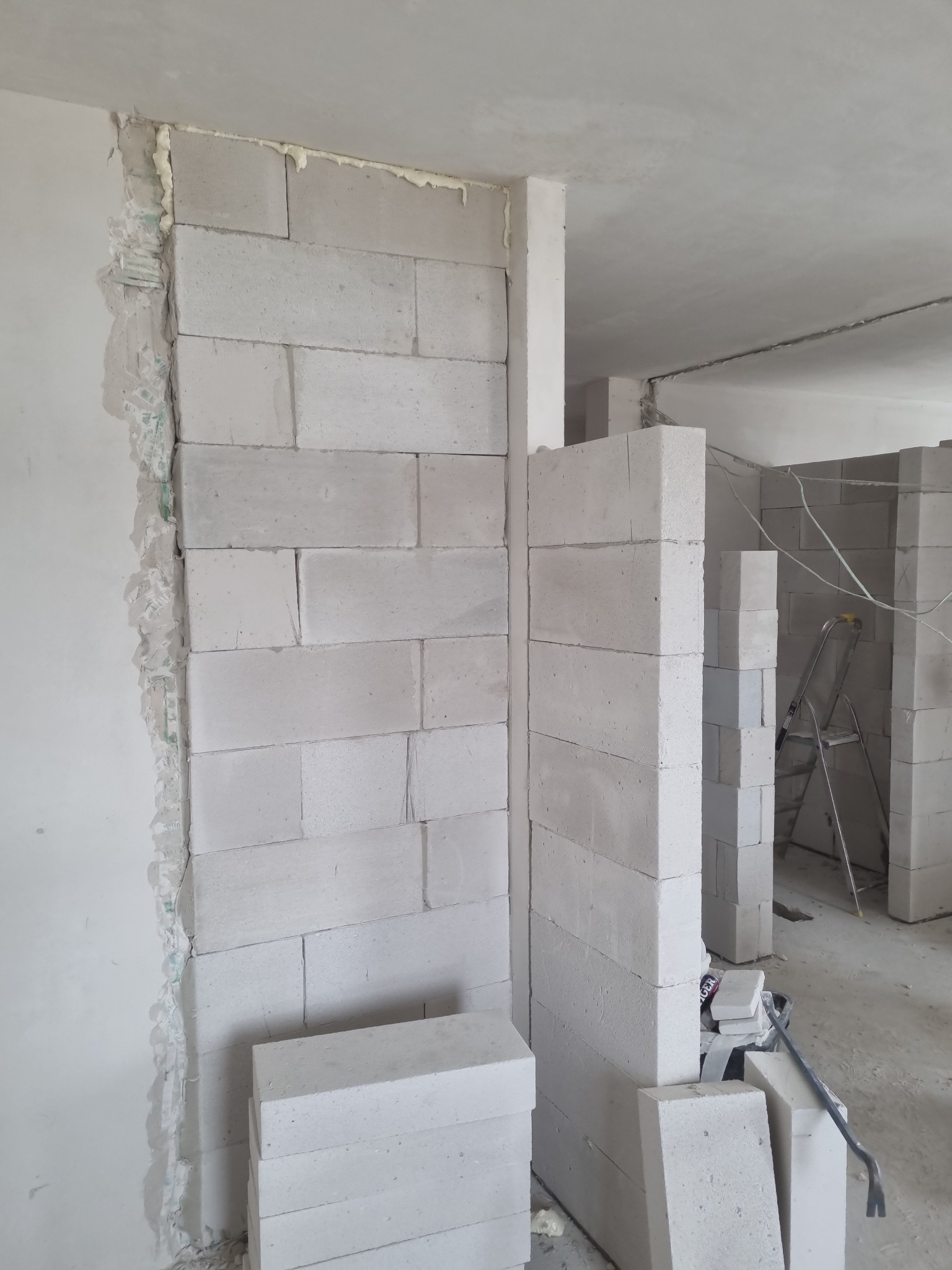 Wznoszenie ściany działowej z białych bloczków betonowych, widoczne spoiny wypełnione pianką montażową, surowe wnętrze w trakcie budowy, drabina w tle.