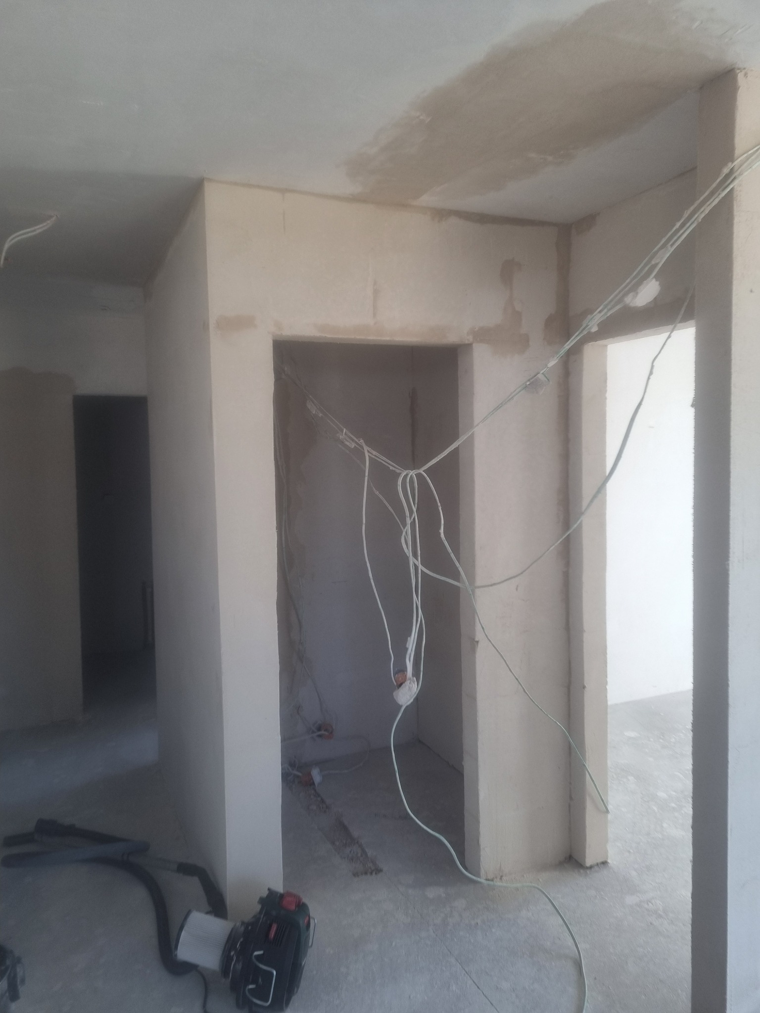 Wnętrze w trakcie remontu: surowe ściany po tynkowaniu, widoczne przewody elektryczne i odkurzacz przemysłowy na podłodze.