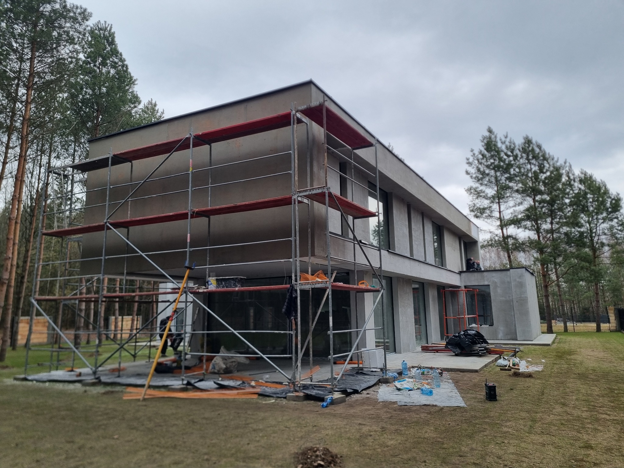Nowoczesny dom w trakcie wykończenia elewacji, z widocznym rusztowaniem, elementami ocieplenia i pracownikiem na dachu, otoczony drzewami i trawnikiem.