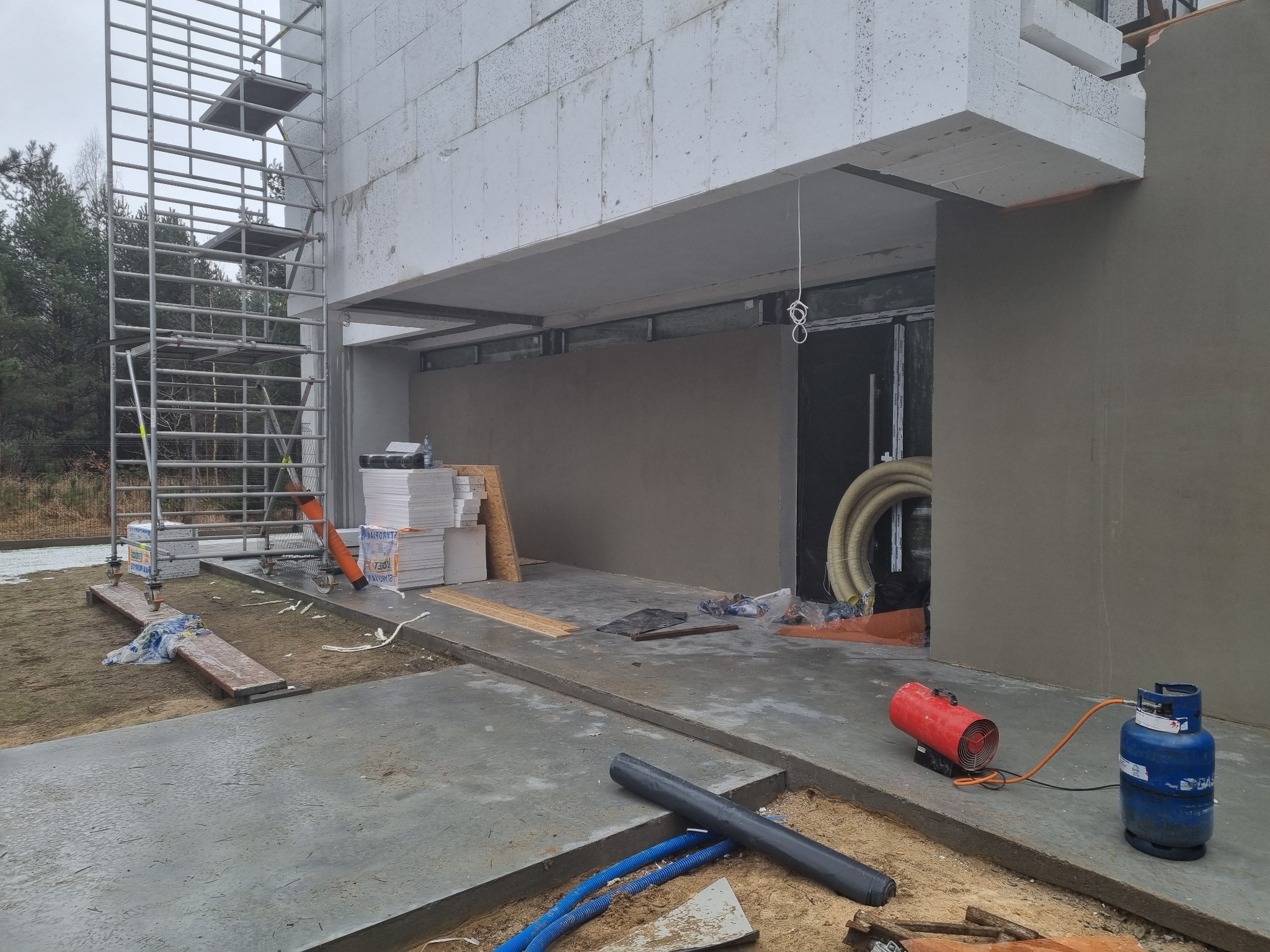 Budowa domu w trakcie: ściany z betonu komórkowego, rusztowanie, otynkowana elewacja, przewody wentylacyjne, nagrzewnica gazowa i butla, widoczne materiały izolacyjne.