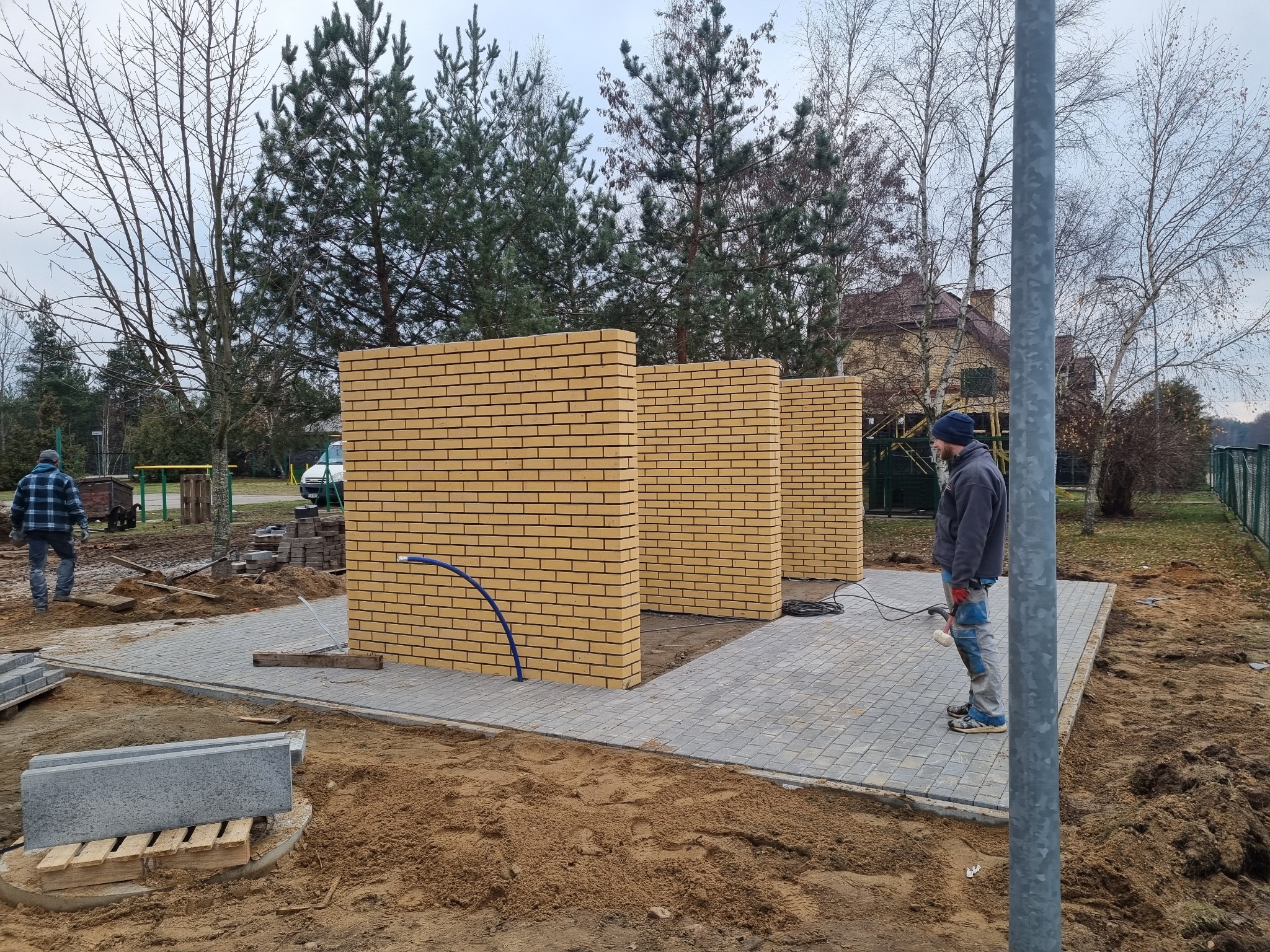 Wznoszenie ścianek działowych z żółtej cegły klinkierowej na zewnątrz budynku, widoczni dwaj pracownicy w trakcie prac wykończeniowych, teren przygotowany pod dalsze prace.