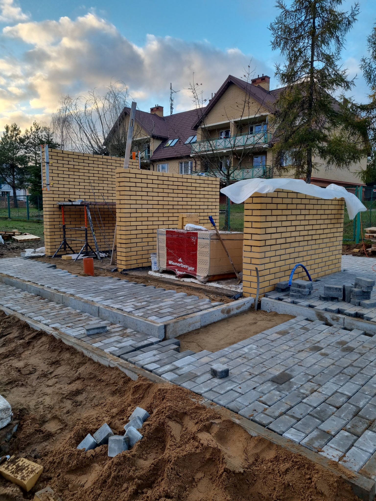 Wznoszenie ścian z żółtej cegły klinkierowej na budowie, widoczne rusztowanie, paleta cegieł Wienerberger i częściowo ułożona kostka brukowa na piasku, tło stanowi dwupiętrowy budynek mieszkalny.