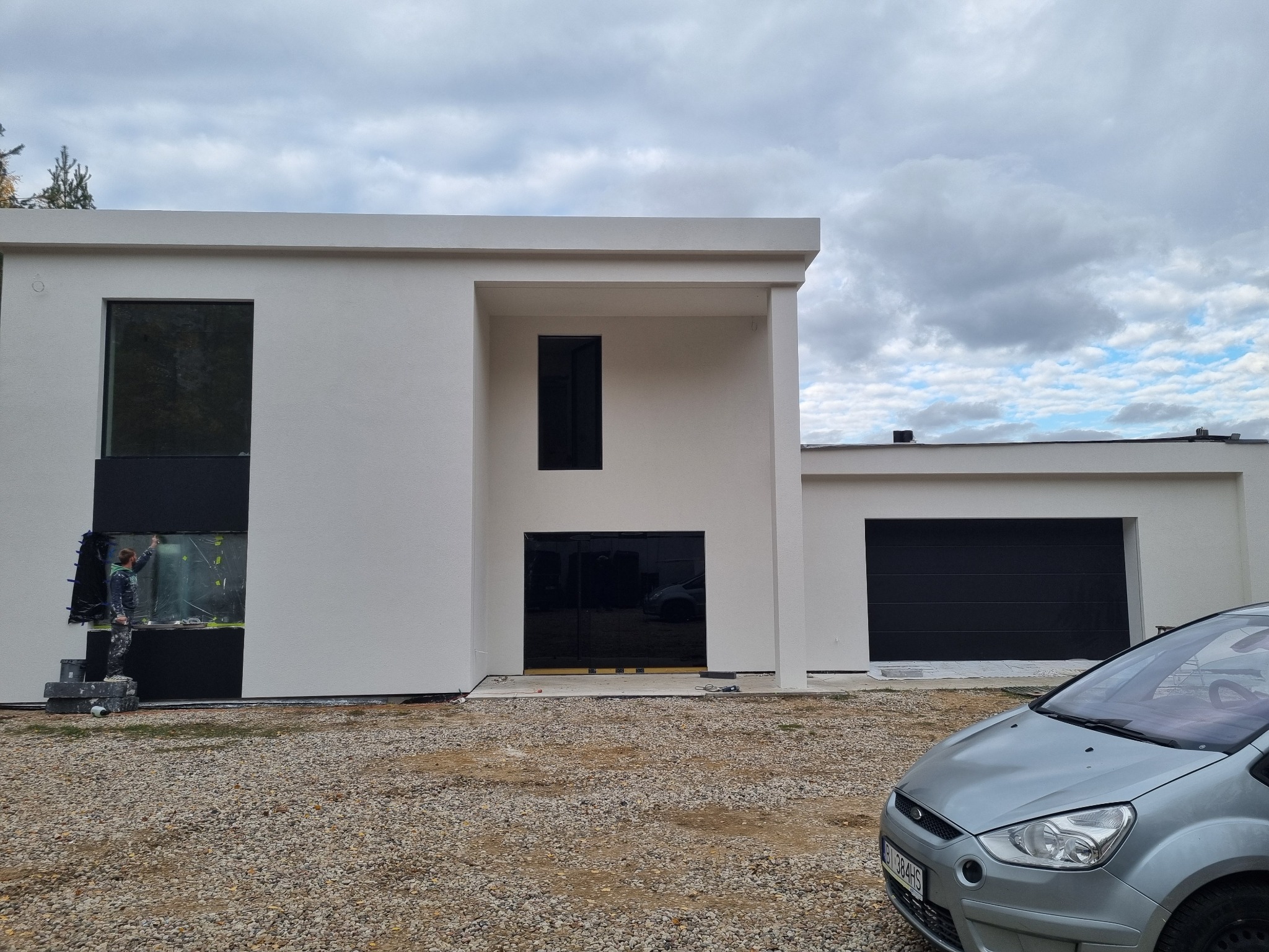 Nowoczesny dom z białymi ścianami i czarnymi oknami, pracownik na rusztowaniu maluje elewację w pobliżu okna, widoczny samochód na żwirowym podłożu.
