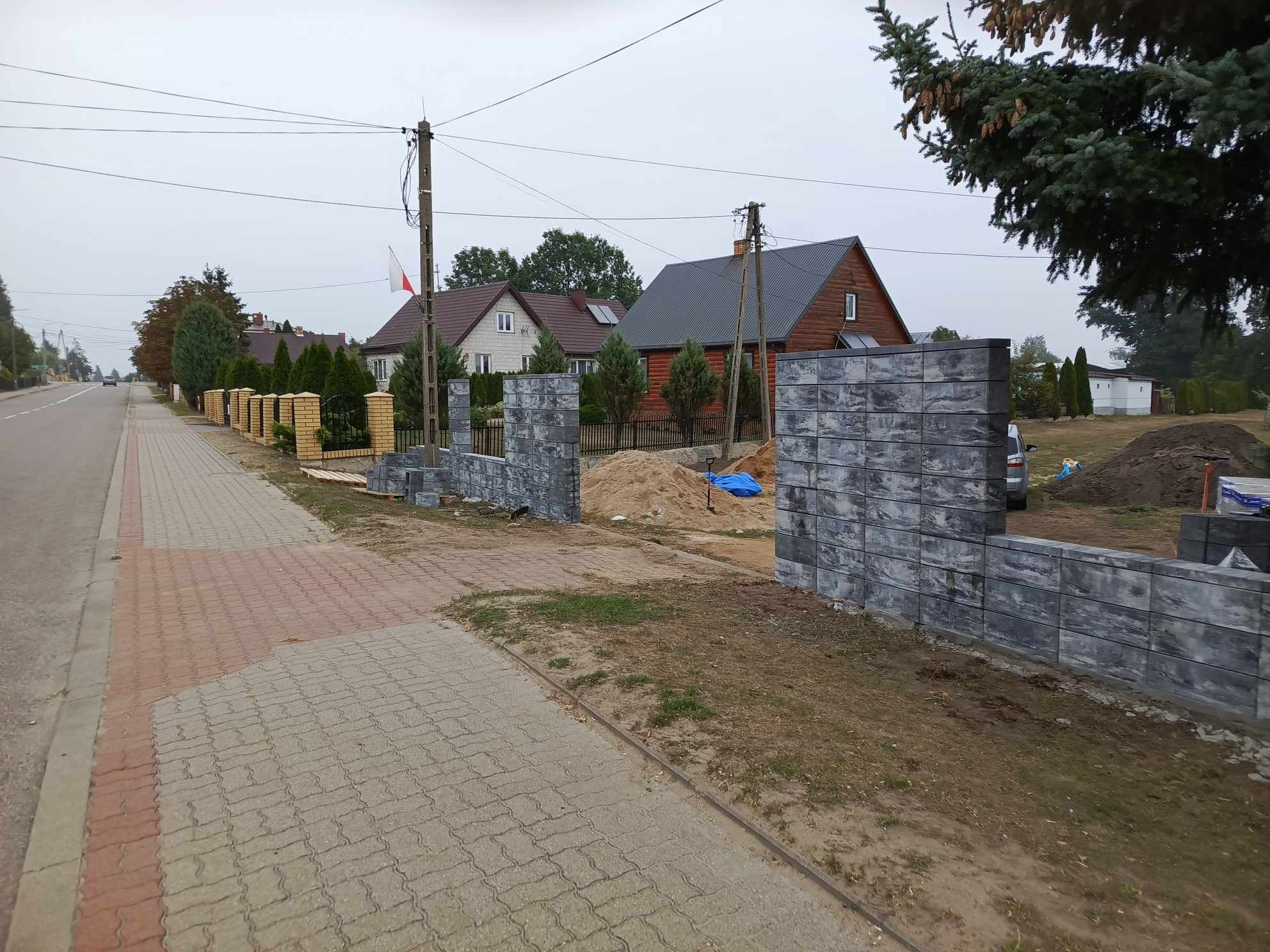 Wznoszenie ogrodzenia z szarej, betonowej kostki modułowej w sąsiedztwie chodnika z kostki brukowej i drogi, widoczne domy w tle.