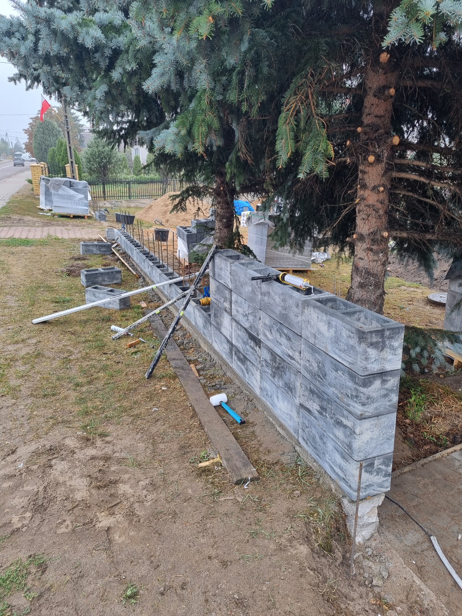 Wznoszenie muru z szarych betonowych bloczków w ogrodzie, widoczne narzędzia: poziomica, młotek gumowy, pistolet do kleju, w tle drzewa iglaste i palety z bloczkami.